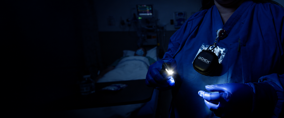 Healthcare worker in blue scrubs wearing an illuminated DEX FingerLight over a blue glove, with a DEX carrying case clipped to their badge.